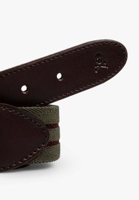 Correa de cinturón de cuero marrón oscuro con una sección de tela verde que presenta rayas rojas y un logo de calavera y huesos cruzados grabado en el lazo.