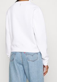 Sweatshirt blanc avec un col rond, des poignets et un ourlet côtelés. Présente un petit symbole gris à l'arrière. Associé à un jean bleu clair.