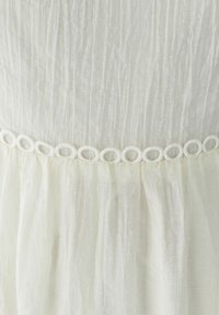 Witte geplooide stof met een gestructureerd uiterlijk, voorzien van een decoratieve lusvormige rand bij de taille. Zacht en licht materiaal.