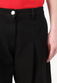 Zwarte high-waisted broek met plooien, voorzien van een zilveren drukknopsluiting. Gemaakt van een gestructureerde stof, met rode draadaccenten.