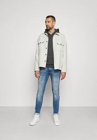 Chaqueta gris claro sobre una sudadera con capucha oscura, combinada con jeans azules y zapatillas blancas. La chaqueta tiene dos bolsillos delanteros y cierre de botones.