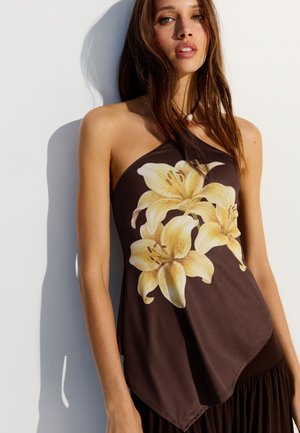 Giovane donna con lunghi capelli castani che indossa un top marrone con grandi stampe floreali di gigli gialli, in piedi contro uno sfondo bianco semplice.