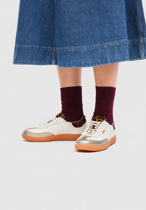 Gambe con gonna di jeans, calzini color granata e scarpe da ginnastica bianche con dettagli leopardati e suole arancioni su uno sfondo chiaro.