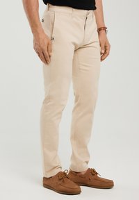 Férfi bézs színű, slim fit nadrágban és barna bőr vitorláscipőben, állva, kezeivel lazán oldalán, egyszerű háttér előtt.