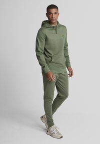 Olivgrüner Hoodie und passende Jogginghose aus glattem Stoff. Der Hoodie hat eine Fronttasche und eine verstellbare Kordel. Mit gelbem Logo akzentuiert.