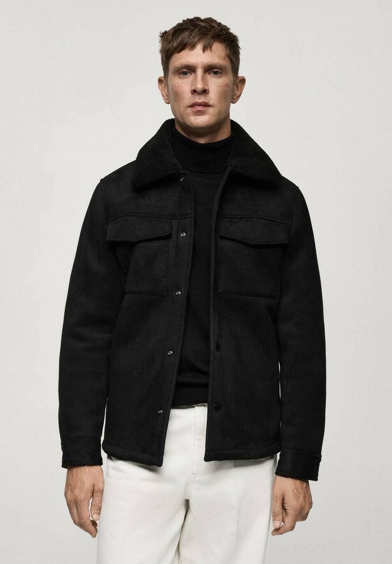 Giacca in suede nera con colletto in Sherpa, due tasche sul petto e bottoni a pressione, abbinata a un dolcevita nero e pantaloni bianchi.