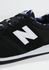 Chaussure de sport noire avec une tige en tissu texturé, logo "N" blanc et lacets noirs. Doublure intérieure avec motif bleu et violet. Semelle en caoutchouc.
