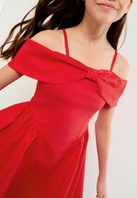 Rød kjole med off-the-shoulder ærmer, der har en stor sløjfe detalje ved brystet og en flagrende nederdel med sammenfoldet tekstur.