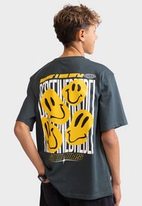 Mörkgrön t-shirt med korta ärmar som har gula smiley-ansikten och grafisk text på baksidan. Tillverkad av bomull, avslappnad passform, rundad halsringning.