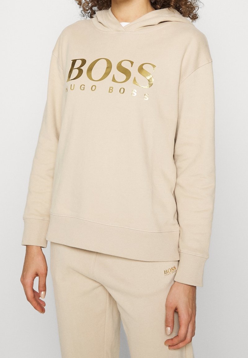 Personne portant un ensemble beige sweat à capuche et pantalon avec le logo doré "BOSS HUGO BOSS" sur la poitrine et le pantalon.