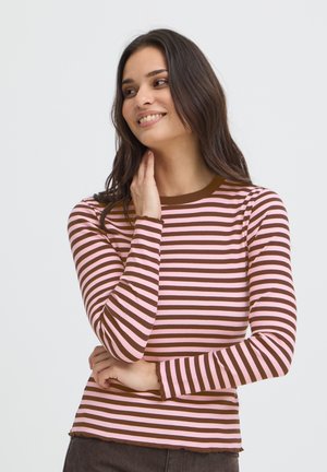 Jeune femme souriante regardant sur le côté, portant un t-shirt à manches longues rayé horizontalement rose et marron avec un pantalon foncé.