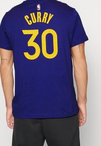 Fialové tričko s nápisem "CURRY" nad číslem "30" ve žluté barvě. Má kulatý výstřih a krátké rukávy, vyrobené z měkké tkaniny.