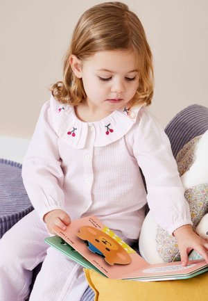 Ungt barn i lyserøde pyjamas med kirsebærkrave, der sidder på en pude, kigger ned og rører ved en farverig interaktiv billedbog.