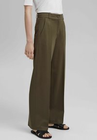 Femme portant un pantalon large taille haute vert olive avec poches, associé à un haut blanc et des sandales noires à bouts ouverts.
