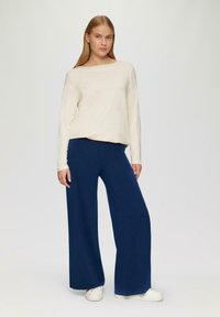 Crème lange mouwtop van zachte stof gecombineerd met wijduitlopende marineblauwe broek van een gestructureerd, lichtgewicht materiaal. Witte sneakers maken de outfit compleet.