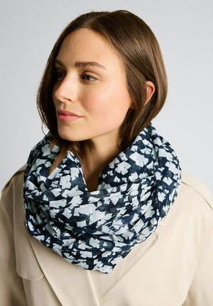 Frau mit kurzen braunen Haaren, die einen beigen Mantel und einen schwarz-weiß geblümten Schal um den Hals trägt.