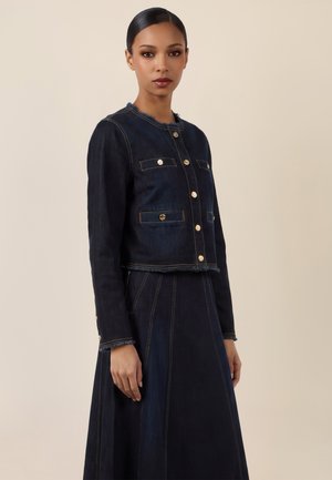 Frau trägt eine dunkelblaue Jeansjacke mit goldenen Knöpfen und passenden, hoch taillierten Rock, steht vor einem beigen Hintergrund.