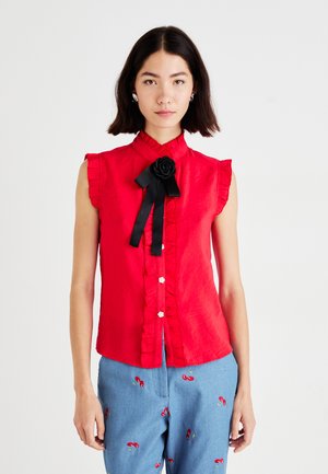 Rød ermeløs bluse med volangkant, frontknapplukking og en svart roseaccent bundet med et satengbånd. Glatt tekstur.