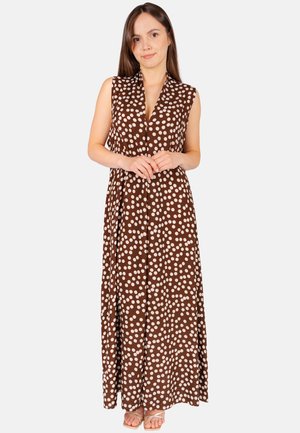 Frau in einem ärmellosen braunen Maxikleid mit weißen Punkten, stehend mit verschränkten Händen und offenen Sandalen.