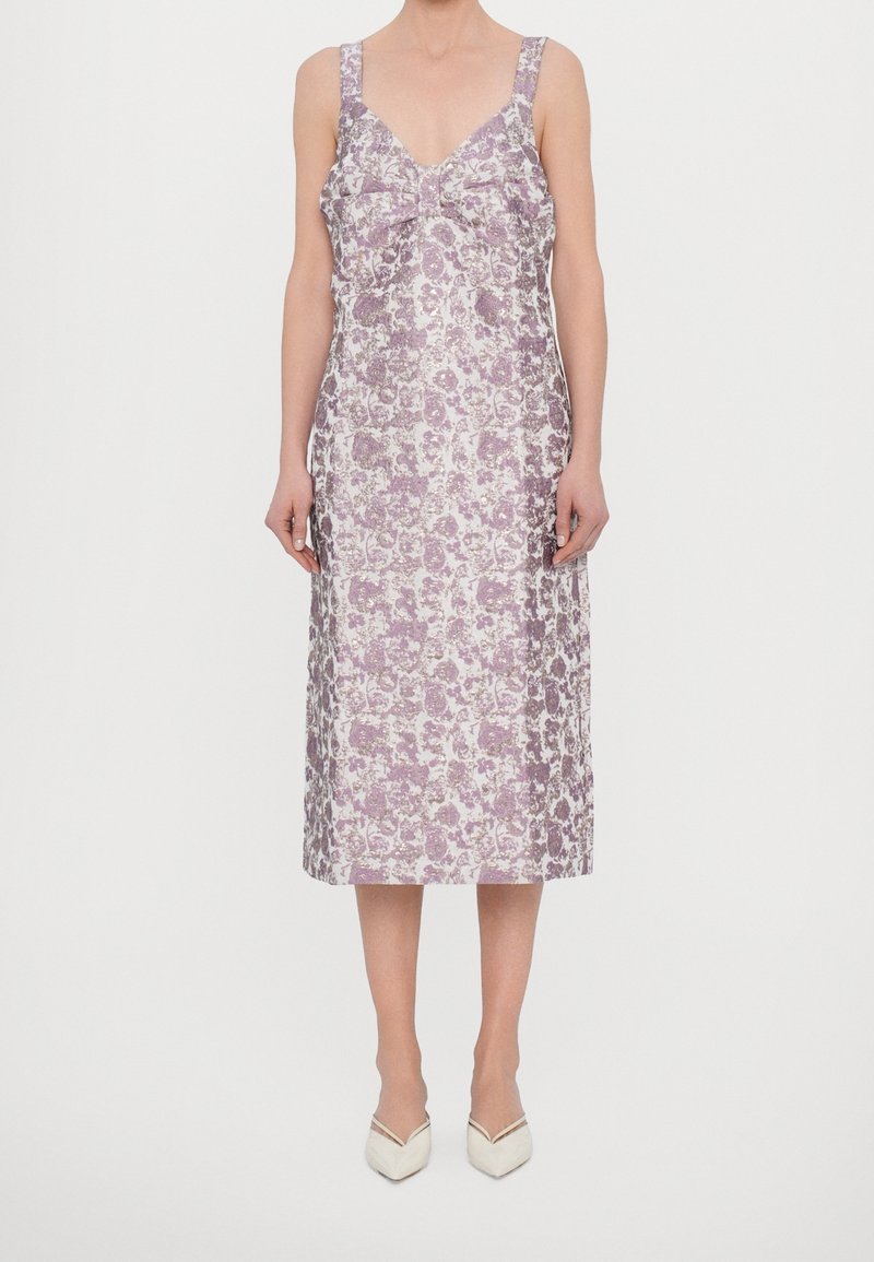 Femme portant une robe sans manches longueur genou avec un motif floral rose et blanc et des chaussures blanches à bouts pointus, debout devant un fond uni.