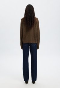 Brūns adījuma džemperis ar brīvu piegriezumu, apvienots ar tumši ziliem taisnajiem džinsiem. Džemperim ir nedaudz īsāka apakšdaļa un garas piedurknes.