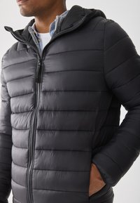 Hombre con una chaqueta acolchada negra con cremallera y capucha, con la mano en el bolsillo, superpuesta a una camisa azul clara y un suéter gris.