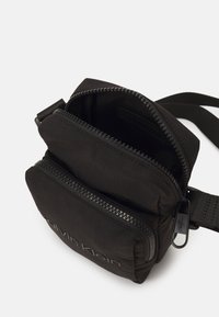 Borsa a tracolla nera in nylon con doppie tasche con zip, texture morbida e logo Calvin Klein in rilievo sulla parte anteriore. Include una tracolla regolabile.