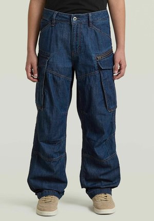 Persoon draagt losse donkerblauwe denim cargobroek met meerdere zakken en beige sneakers, staand tegen een effen achtergrond.