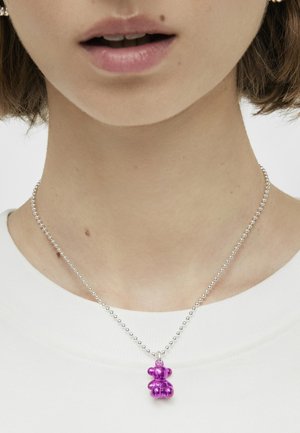 Collar de cadena de bola plateada con un colgante en forma de oso morado. El colgante es brillante, compacto y se muestra de manera prominente sobre una camiseta blanca lisa.
