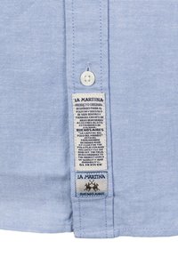 Vaaleansininen nappipaita, jossa on kudottu rakenne. Sivussa on hoito-ohjetarra, jossa on merkki- ja sijaintitiedot.