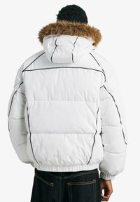 Biela prešívaná bunda s hnedou kapucňou lemovanou kožušinou, s čiernymi dekoratívnymi líniami, elastickými manžetami a hranatým strihom.