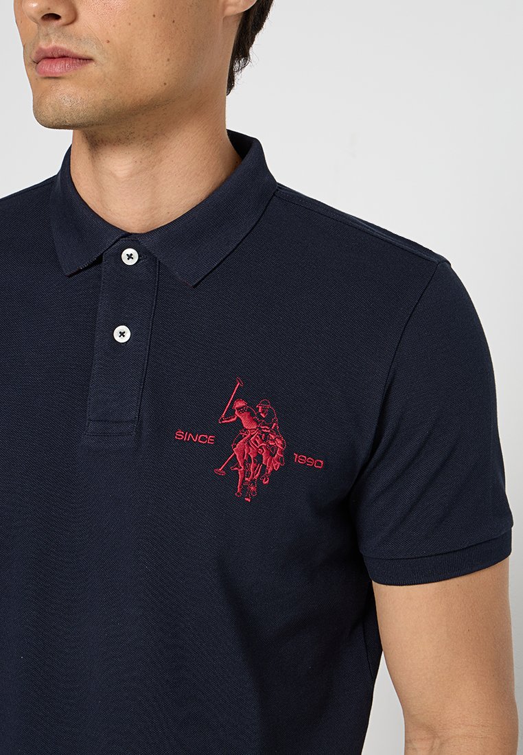 Polo shirt marine en tissu texturé avec un logo rouge brodé. Col à trois boutons avec boutons blancs ; détail "DEPUIS 1990" inclus.