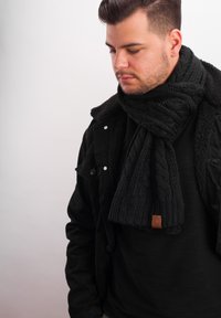 Black knitted scarf with cable pattern and ribbed texture, featuring a small leather tag. Worn over a black sweater and jacket.