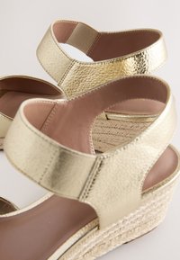 Sandales compensées métalliques dorées avec des lanières texturées, un talon fermé et une semelle intérieure beige. Base enveloppée de jute offrant une hauteur supplémentaire et du détail.