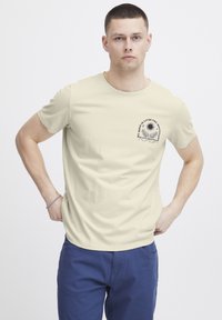 Kortermet, lys beige t-skjorte med rund hals. Har et trykt design på venstre bryst, laget av myk, pustende stoff.