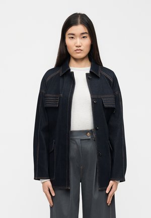 Tânără cu păr lung și închis la culoare, purtând o jachetă de denim neagră peste un pulover alb și pantaloni gri cu talie înaltă, stând în fața unui fundal simplu.