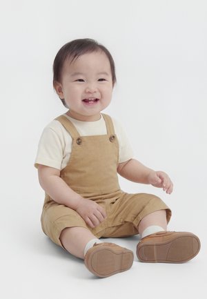 Lächendes Kleinkind sitzt auf dem Boden, trägt beige Latzhose, weißes Hemd, weiße Socken und beige Schuhe, vor einem schlichten hellen Hintergrund.