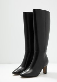 Bottes noires en cuir montant jusqu'au genou avec un bout pointu et un talon empilé moyen, présentant une texture lisse et un design minimaliste.