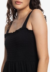 Ung kvinna med långt mörkt lockigt hår som bär en svart ärmlös klänning med smockad liv och volang i halsen.