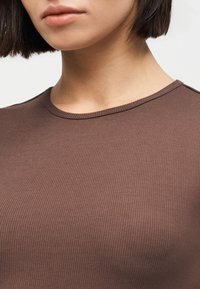 Κοντινό πλάνο ενός ατόμου που φοράει καφέ ριμπ μπλούζα με στρογγυλή λαιμόκοψη, με κοντά σκουρόχρωμα μαλλιά και ουδέτερο χρώμα στα χείλη.