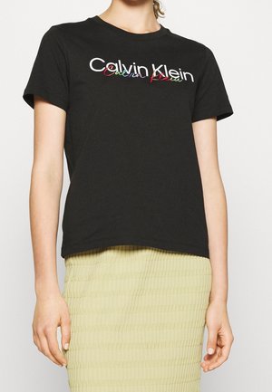 Czarny, krótki t-shirt z białym i wielokolorowym logo "Calvin Klein", noszony z beżową, teksturowaną spódnicą, pokazany od szyi do połowy uda.