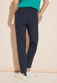 Pantaloni su misura blu navy in tessuto liscio, con vita elastica e due tasche posteriori, abbinati a sneakers bianche, visti da dietro.