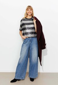 Haut court à rayures noires et argentées pailletées, jean large en denim bleu, et un manteau bordeaux porté sur un bras, assorti à des bottines noires.