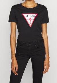 Kvinde iført en sort Guess t-shirt med et rødt og hvidt trekantet logo samt sorte tætsiddende jeans, stående foran en ensfarvet baggrund.