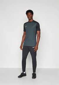 Homem de pé, vestindo uma camisola atlética verde escura e preta, calças de jogging pretas e sapatos de desporto pretos, em fundo branco liso.