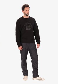 Felpa nera in cotone con una stampa testurizzata e opaca di "DENIM DIVISION 78" al centro, abbinata a jeans in denim scuro e sneakers bianche.