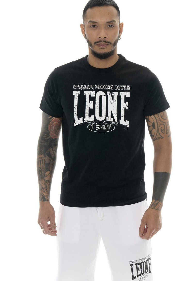 Un mannequin masculin tatoué porte un T-shirt noir de style boxe Leone 1947 et un short blanc sur un fond blanc uni.