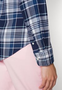 Närbild av en persons arm iklädd en blå och vit rutig skjorta med knäppta manschetter över ljusrosa byxor mot en enfärgad bakgrund.