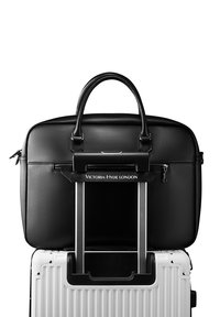 Bolso de portátil de cuero negro con asas dobles, cierre de cremallera y una correa diseñada para ajustarse sobre el mango de una maleta. Presenta acentos de hardware metálico.