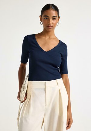 Kvinde iført en mørkeblå V-hals kortærmet top, cremefarvede højttaljede plissee bukser, øreringe i form af ringe, og hun holder en cremefarvet håndtaske.